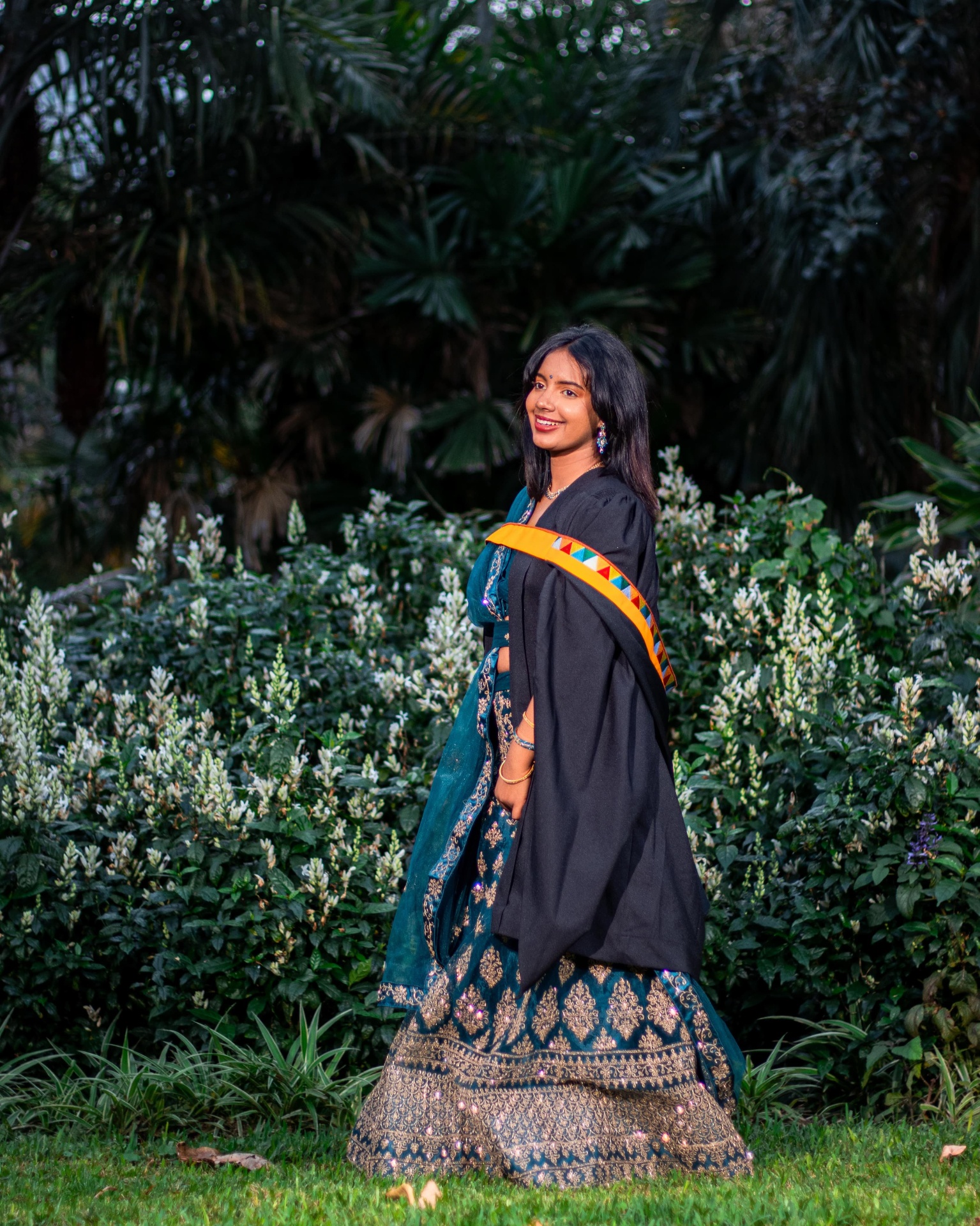 Graduate in a teal lehenga and black gown in a garden