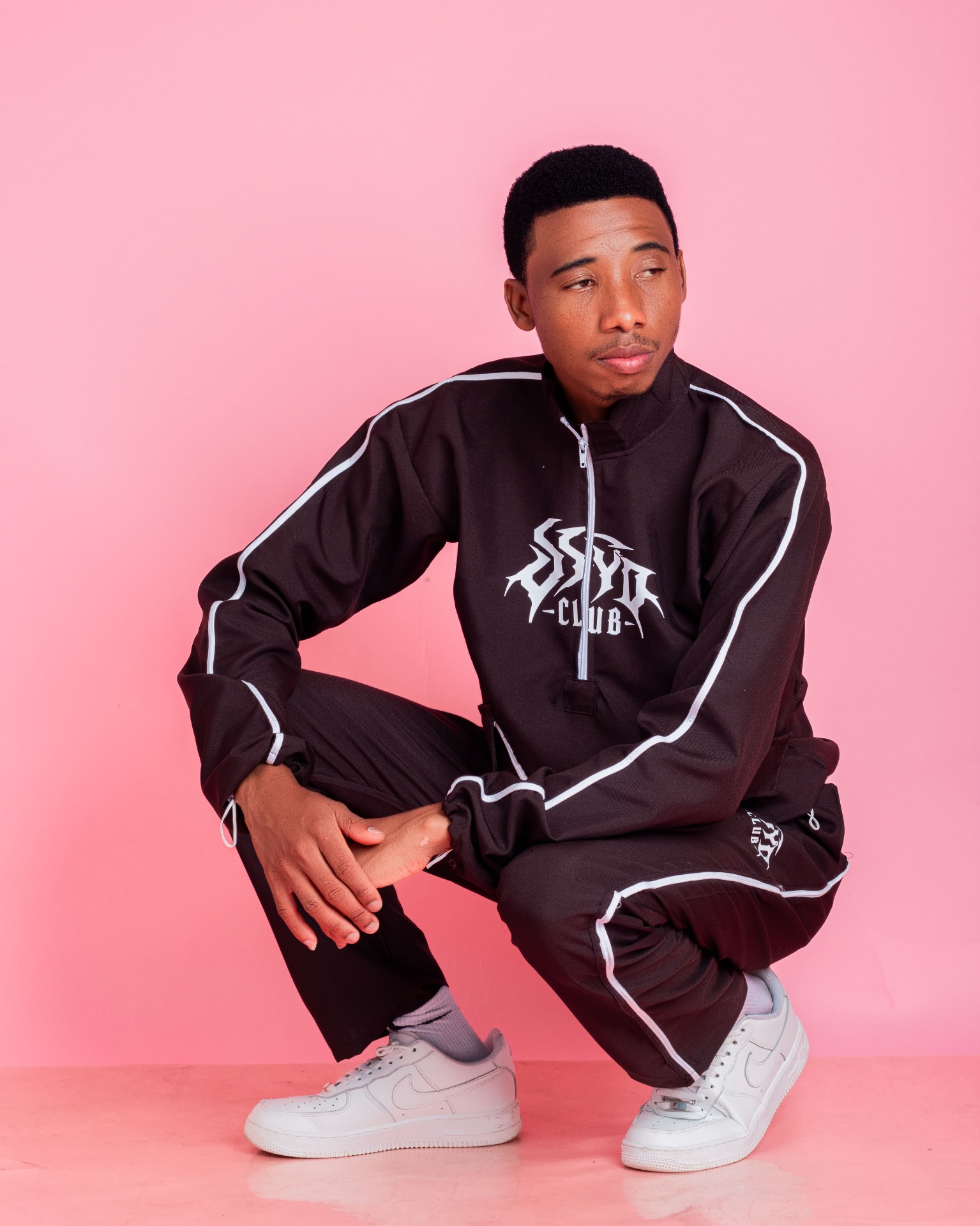 Man crouching in a black tracksuit against a pink backdrop
