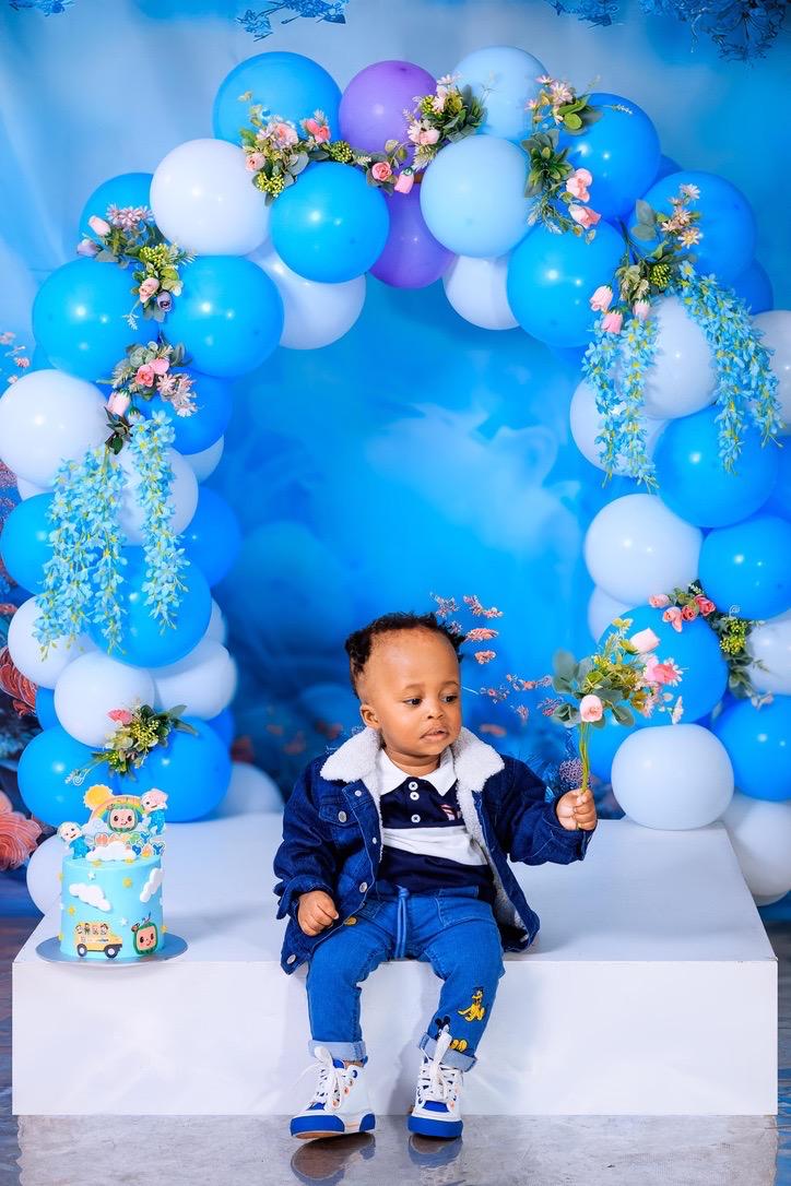 Baby boy in denim holding flowers under a blue balloon arch