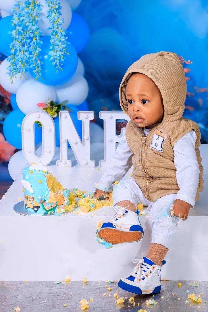 Playful cake smash photo of a one-year-old boy in a beige hoodie