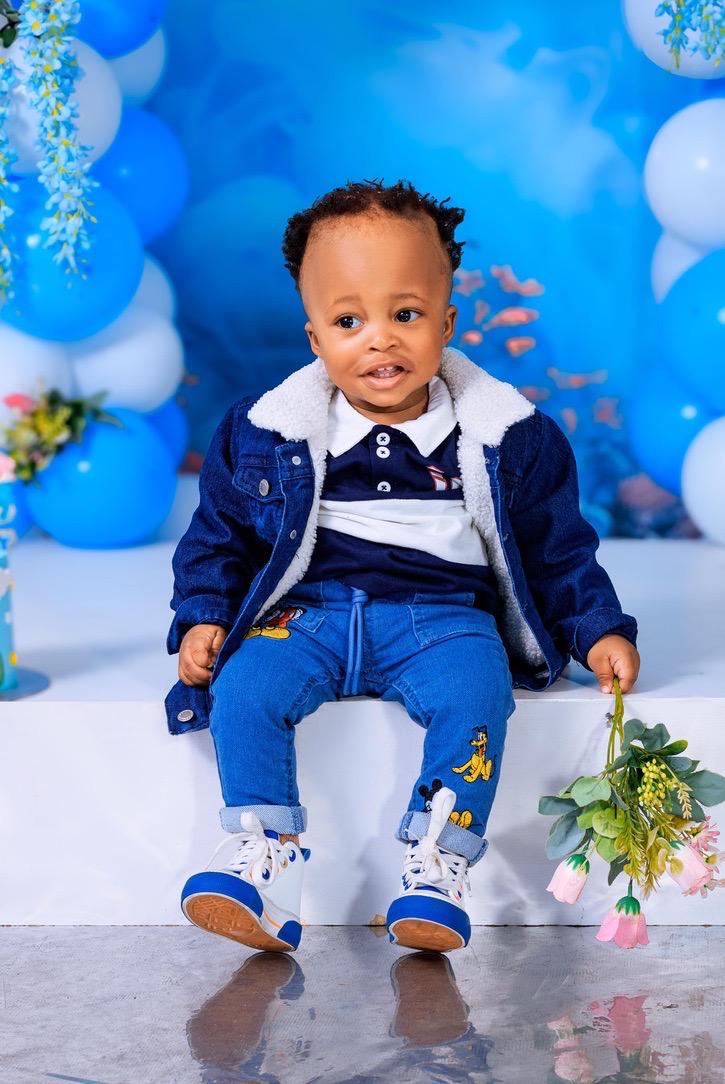 One-year-old boy in a denim jacket seated at a blue balloon arch